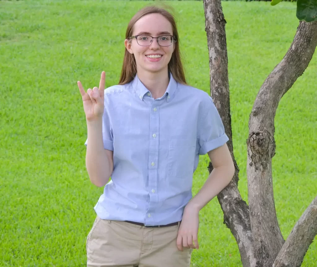 A photo of Olivia from the waist up, smiling and holding up the UT Longhorns sign while leaning on a tree. She is wearing a pastel blue, collared, short sleeve shirt, and khaki pants with a belt.
